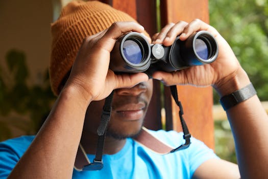 Faceless African American male in beanie looking through binoculars while standing near opened window